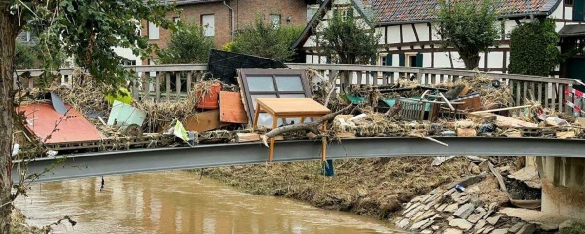 Brücke voller Trümmer nach Hochwasser in einem Dorf, mit Fachwerkhäusern im Hintergrund.