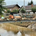 Brücke voller Trümmer nach Hochwasser in einem Dorf, mit Fachwerkhäusern im Hintergrund.