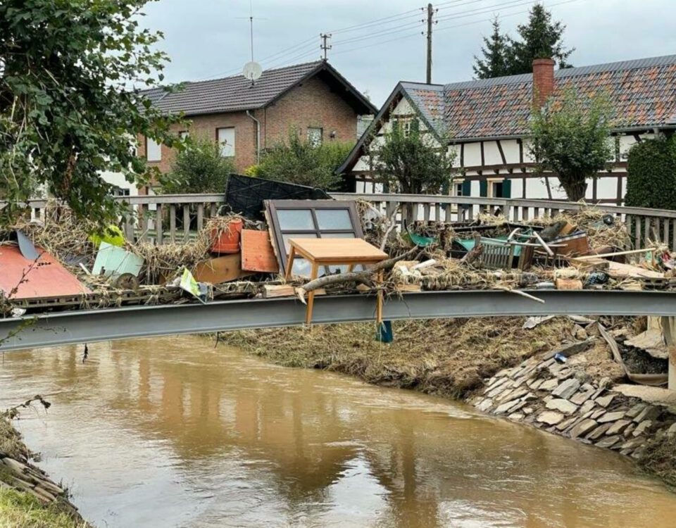 Brücke voller Trümmer nach Hochwasser in einem Dorf, mit Fachwerkhäusern im Hintergrund.
