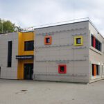 Moderne Schulgebäude-Fassade mit bunten Fenstern und einem Eingangsschild „Heinrich-Heine-Gymnasium“.