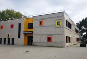 Moderne Schulgebäude-Fassade mit bunten Fenstern und einem Eingangsschild „Heinrich-Heine-Gymnasium“.