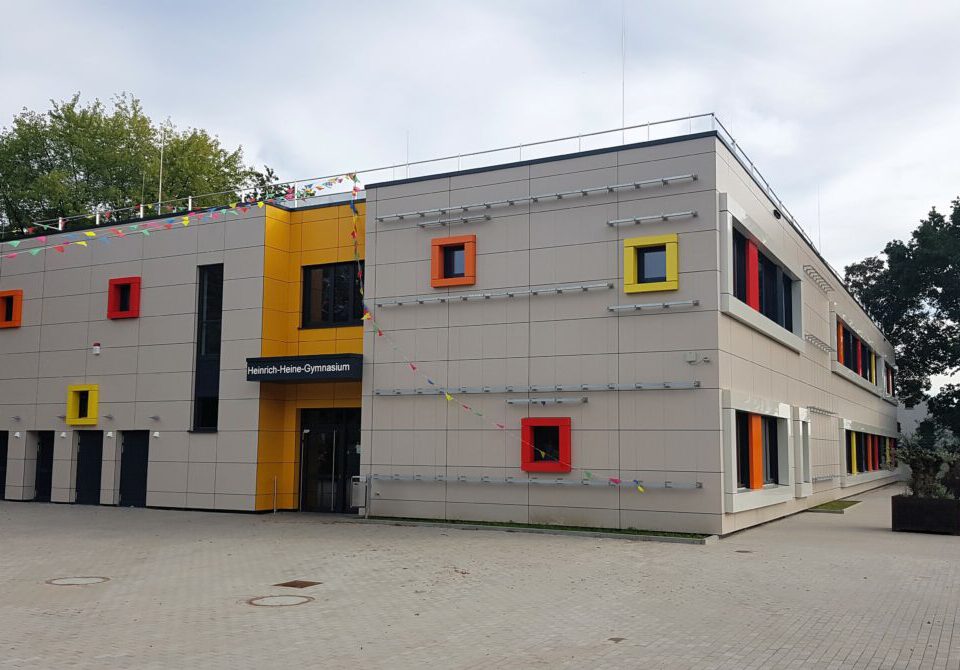 Moderne Schulgebäude-Fassade mit bunten Fenstern und einem Eingangsschild „Heinrich-Heine-Gymnasium“.