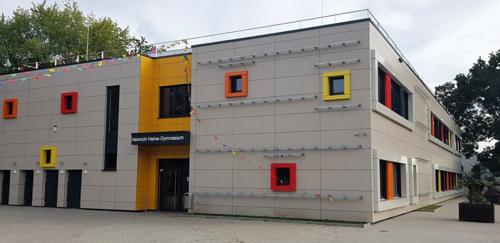 Moderne Schulgebäude-Fassade mit bunten Fenstern und einem Eingangsschild „Heinrich-Heine-Gymnasium“.