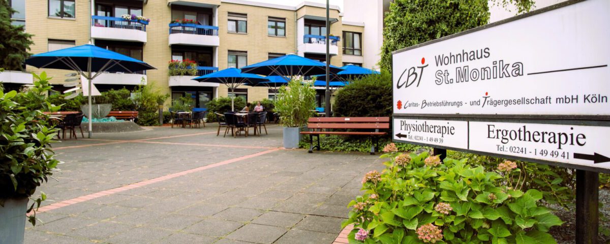 Eingang zum Wohnhaus St. Monika mit Außenbereich und blauen Sonnenschirmen in einem gepflegten Garten.