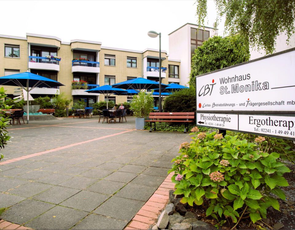 Eingang zum Wohnhaus St. Monika mit Außenbereich und blauen Sonnenschirmen in einem gepflegten Garten.