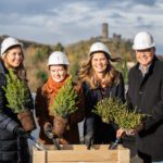 Vier Personen mit Helmen pflanzen Bäume bei einer symbolischen Pflanzzeremonie auf einem Dach mit Natur im Hintergrund.