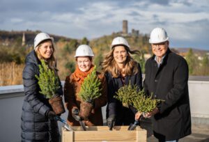 Vier Personen mit Helmen pflanzen Bäume bei einer symbolischen Pflanzzeremonie auf einem Dach mit Natur im Hintergrund.