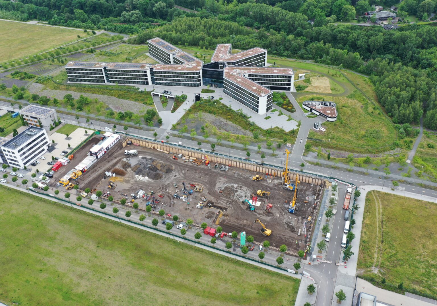 Luftaufnahme einer Baustelle neben einem modernen Bürogebäude, umgeben von grüner Landschaft.