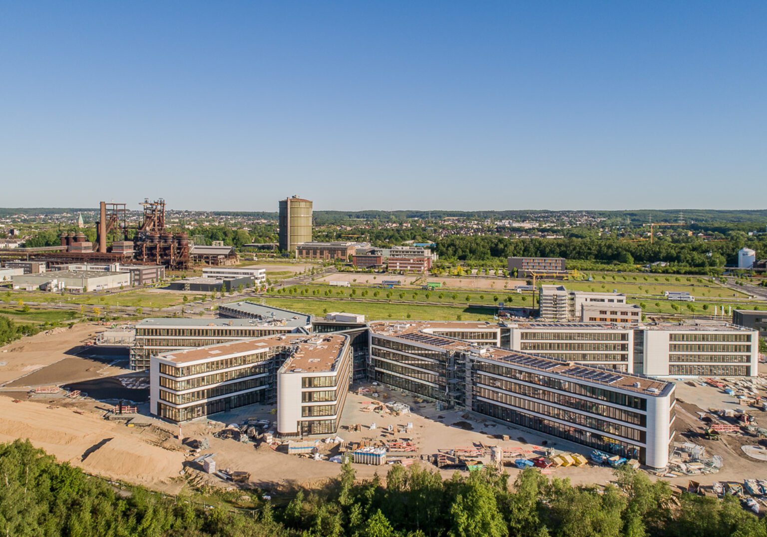 Moderne Bürogebäude im Bau neben einer Industrieanlage, mit grüner Landschaft und blauem Himmel im Hintergrund.