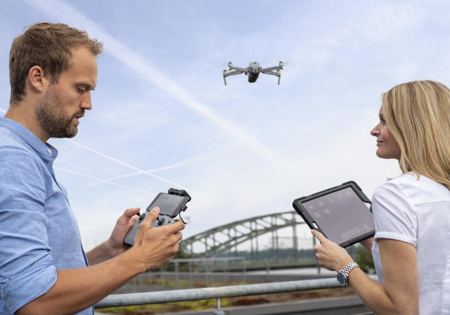Zwei Personen steuern eine Drohne im Freien mit Fernbedienung und Tablet, Brücke im Hintergrund.