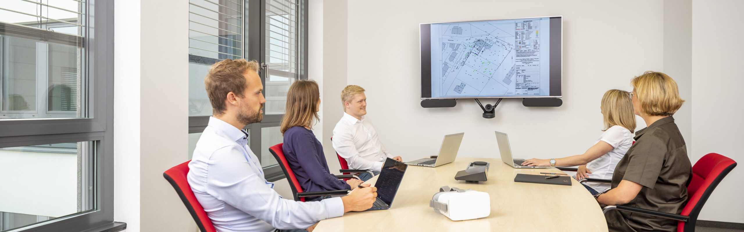 Konferenzraummeeting: Fünf Personen diskutieren über Architekturentwurf auf einem Bildschirm. Laptops auf dem Tisch.