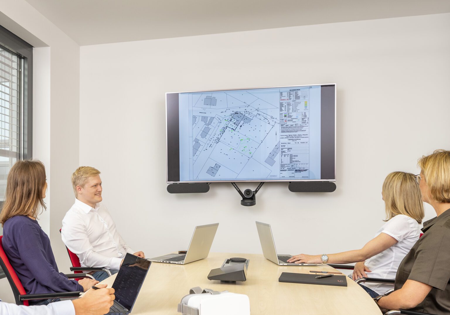 0891_03-1536x1075.2-c-default Menschen in Besprechung mit Bauplan auf Bildschirm, Teamarbeit im modernen Konferenzraum.