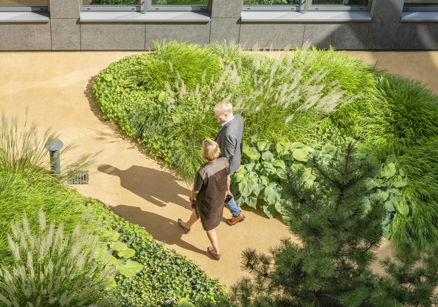 1161_026_AUSWAHL_08-Motiv_Kommunikation-Innenhof-16x5-1-1536x1075.2-c-default Zwei Personen spazieren auf einem Weg durch einen grünen, sonnigen Garten vor einem modernen Gebäude.