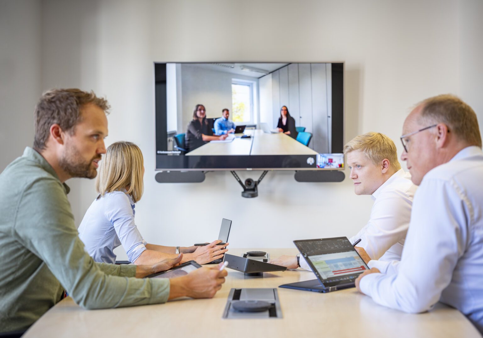 1325_001_AUSWAHL_07-1536x1075.2-c-default Videokonferenz: Fünf Personen im Besprechungsraum diskutieren mit Kolleg*innen auf dem Bildschirm über ein Online-Meeting.