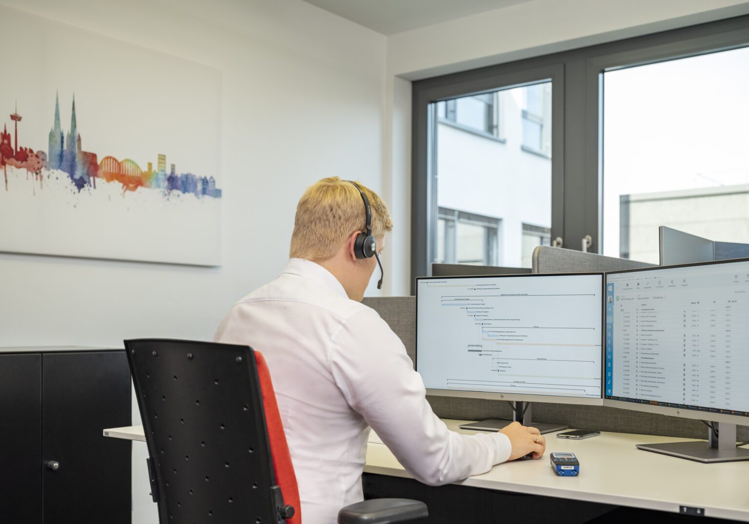 Mann am Schreibtisch mit Headset arbeitet am Dual-Monitor im Büro, Grafik von Köln Skyline an der Wand.
