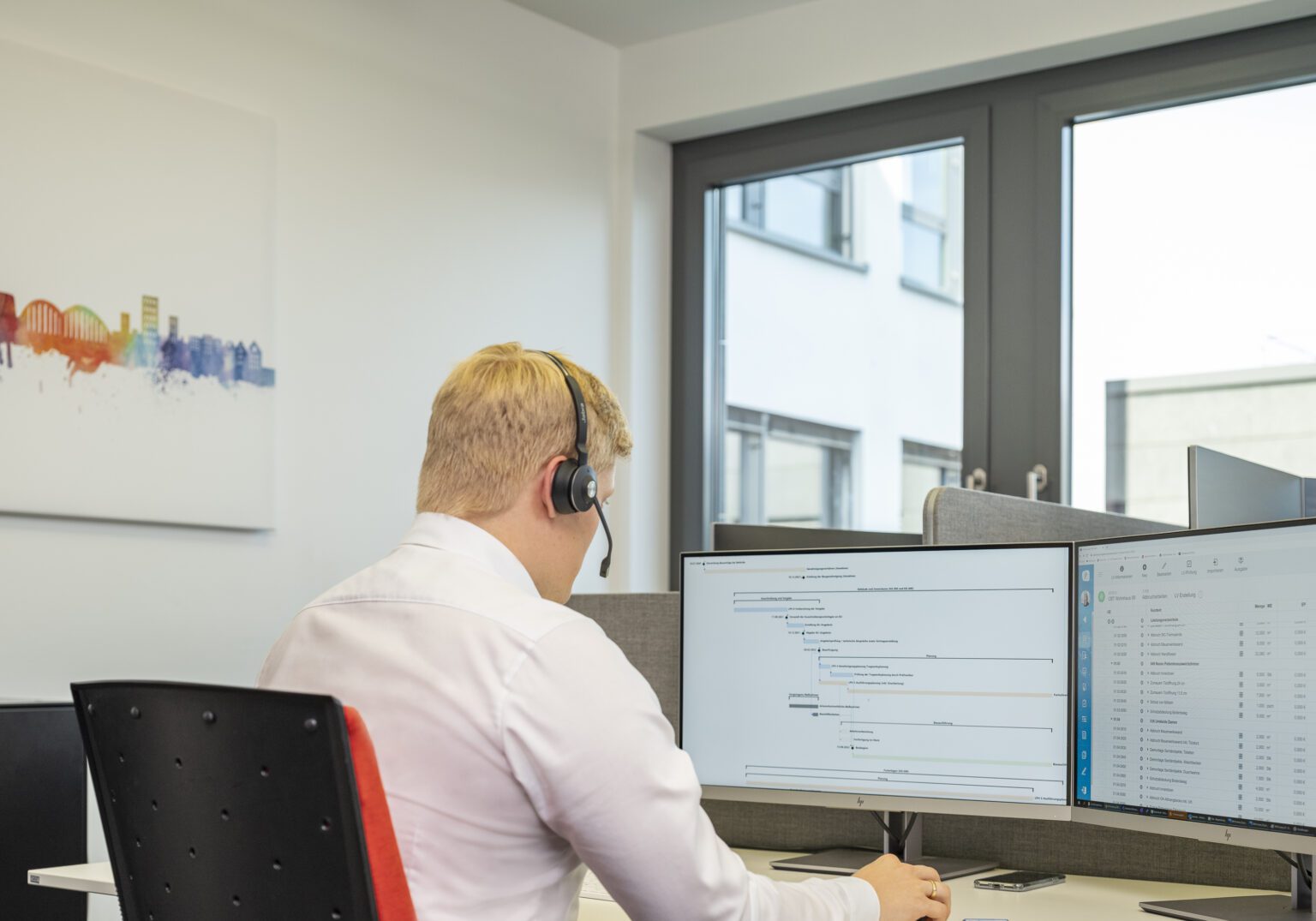 Mitarbeiter am Schreibtisch mit Headset, arbeitet am Computerbildschirm mit Diagramm und Tabellen in modernem Büro.