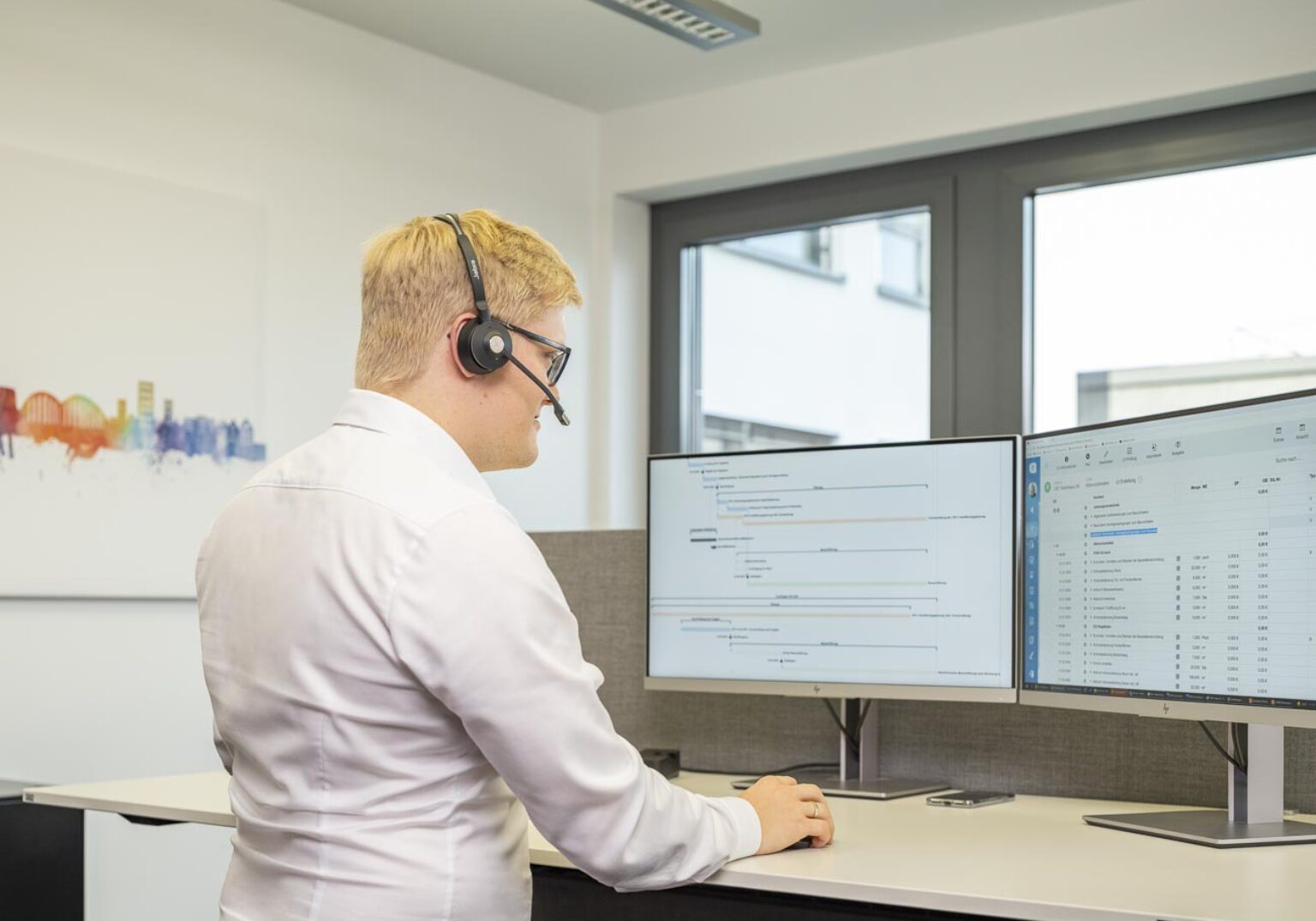 Mann mit Headset arbeitet an zwei Monitoren im Büro, telefoniert und verwendet Softwareanwendungen.