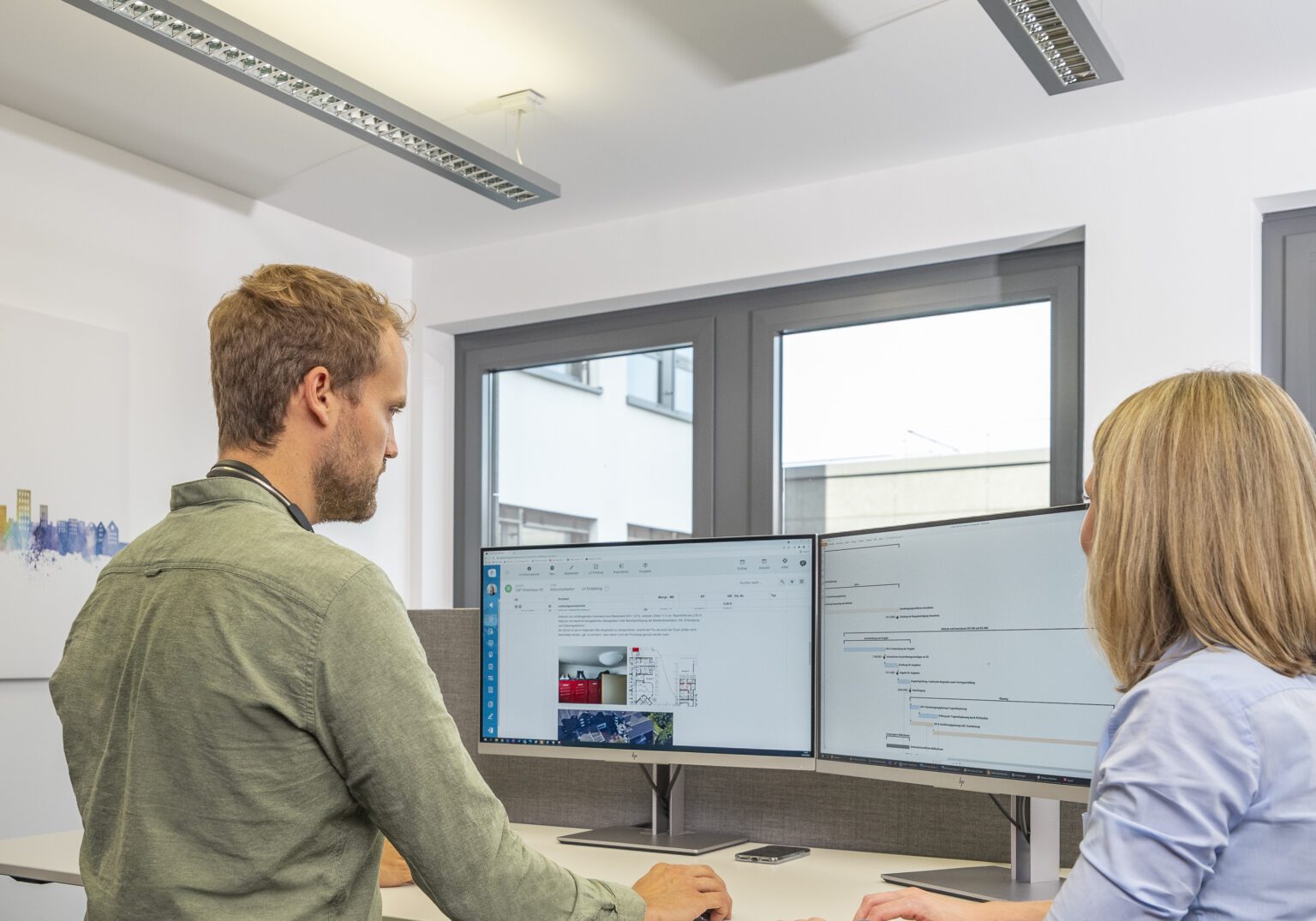 1515_038_AUSWAHL_10-Motiv_Baumanagement-16x5-1-1536x1075.2-c-default Zwei Personen arbeiten im Büro an Computern, besprechen Projekte und analysieren Daten auf Monitoren.