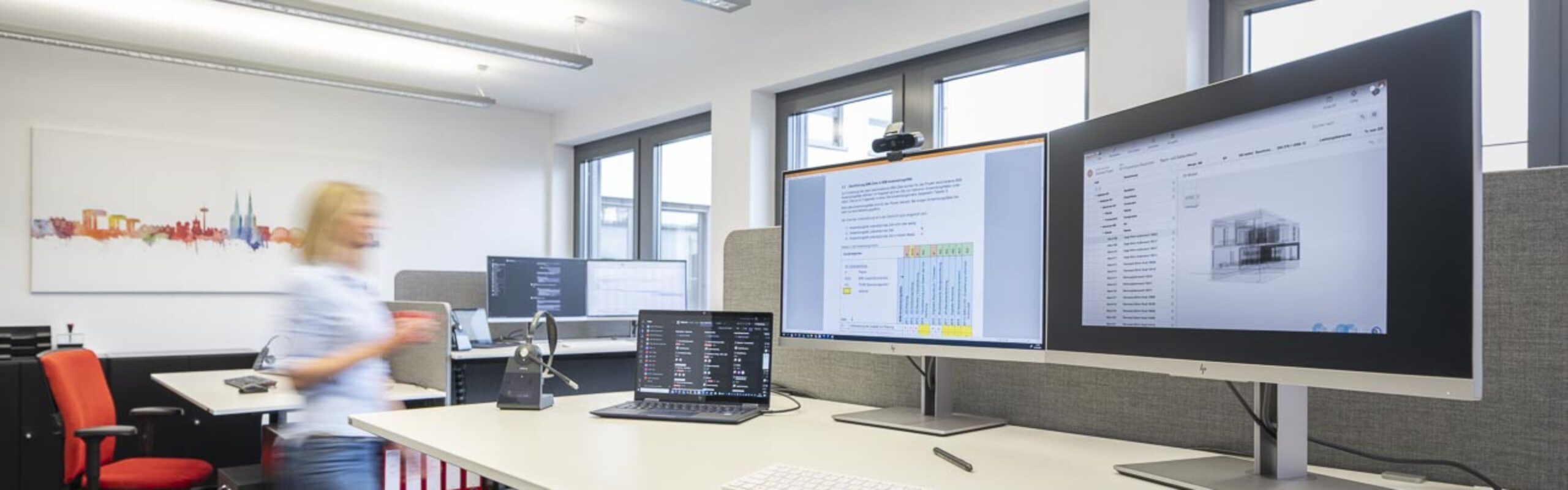 Modernes Büro mit mehreren Monitoren und Laptop auf höhenverstellbarem Schreibtisch, lebendige Wandkunst und ergonomischem Stuhl.