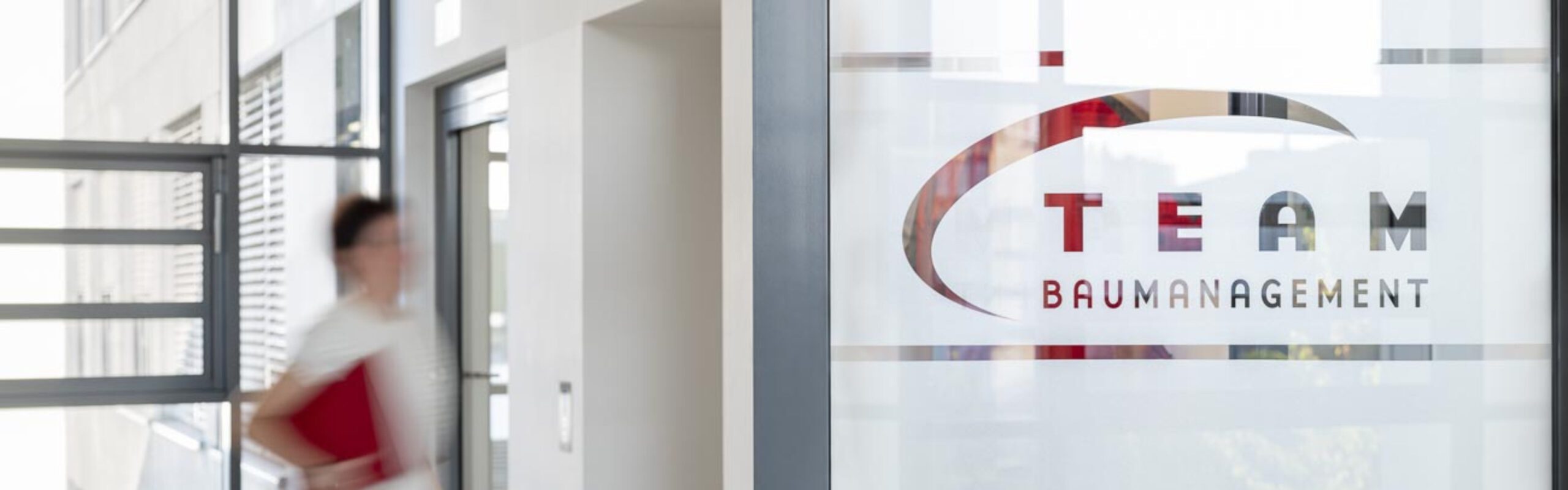 Büroeingang von Team Baumanagement, Frau mit roter Mappe im modernen Bürogebäude.