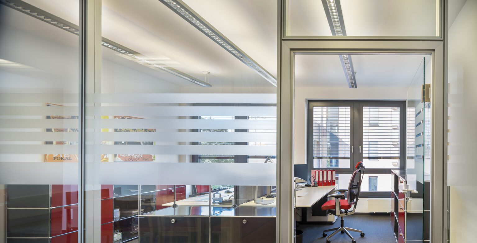 Modernes Büro mit Glaswänden, rotem Bürostuhl und Schreibtisch, das Tageslicht durch Fenster empfängt.