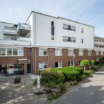 Moderne Wohnanlage mit roter Backsteinfassade und begrüntem Eingangsbereich unter blauem Himmel.