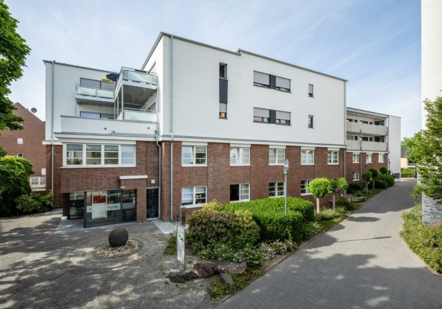 Moderne Wohnanlage mit roter Backsteinfassade und begrüntem Eingangsbereich unter blauem Himmel.