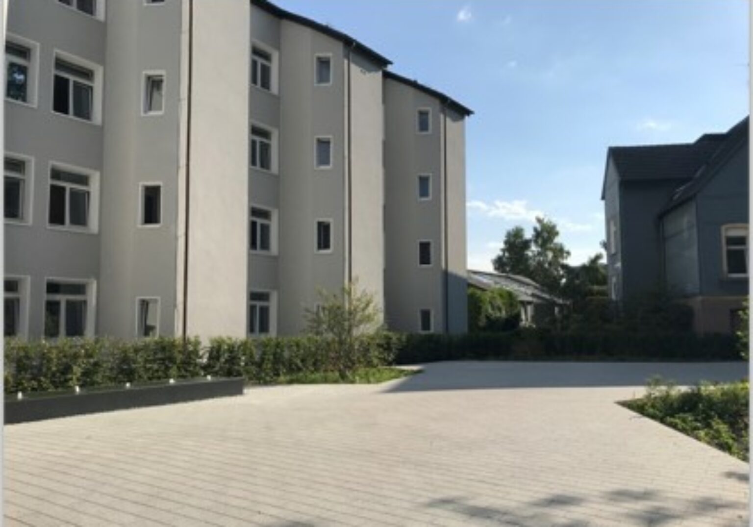 Mehrfamilienhaus mit grauer Fassade, umgeben von gepflegtem Garten und hellgepflasterter Einfahrt unter klarem Himmel.