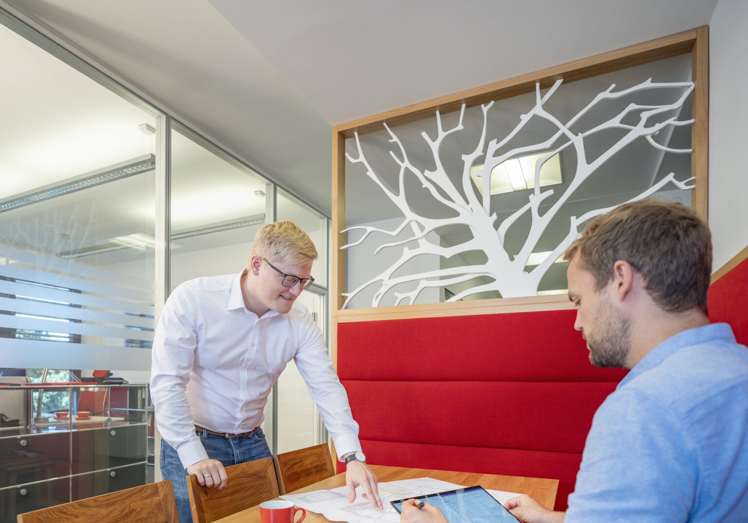 Zwei Männer besprechen Pläne in einem modernen Büro mit rotem Sofa und dekorativer Trennwand.