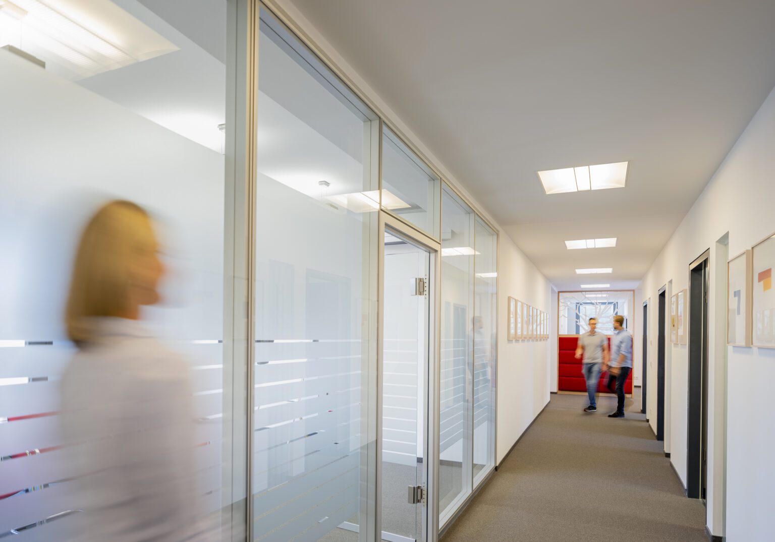 Moderne Büroflur mit verglasten Wänden, unscharfe Personen in Bewegung, helle Beleuchtung.