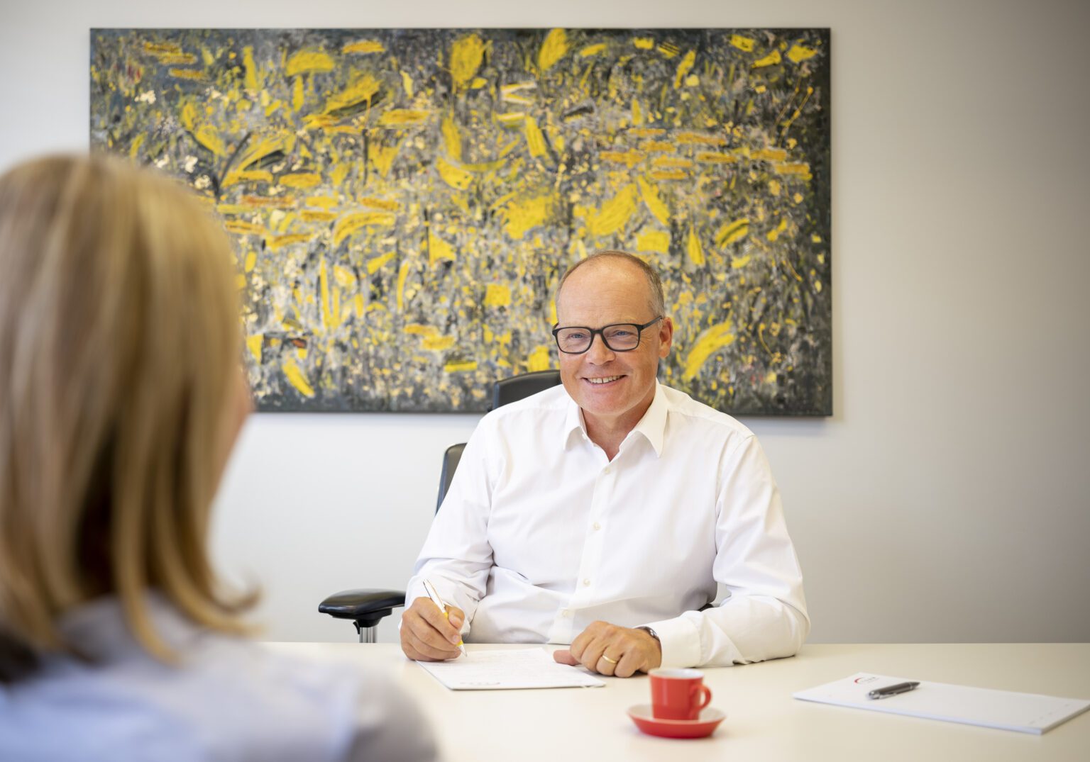 Mann in weißem Hemd beim Meeting, lächelnd, vor abstraktem Gemälde im Büro.