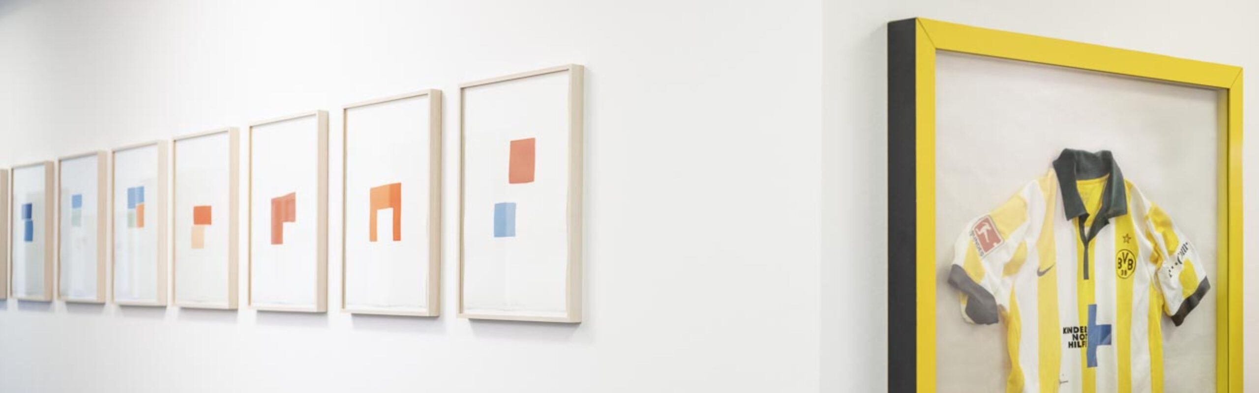 Galeriewand mit abstrakten Kunstwerken und eingerahmtem gelb-schwarzem Fußballtrikot zur Dekoration.