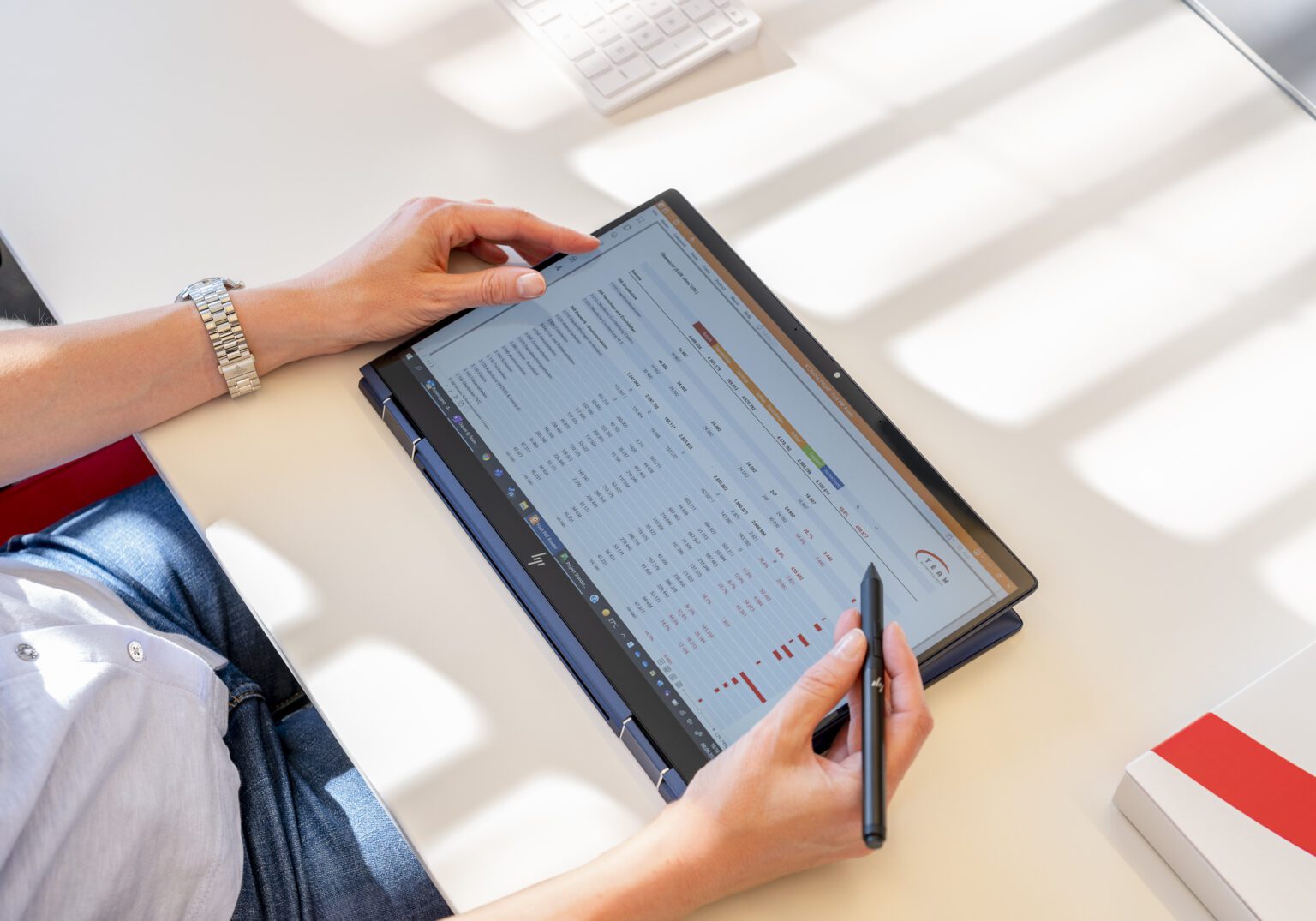 Person bearbeitet Daten auf Tablet mit Stift, zeigt Finanzgrafiken auf Schreibtisch mit Tastatur.