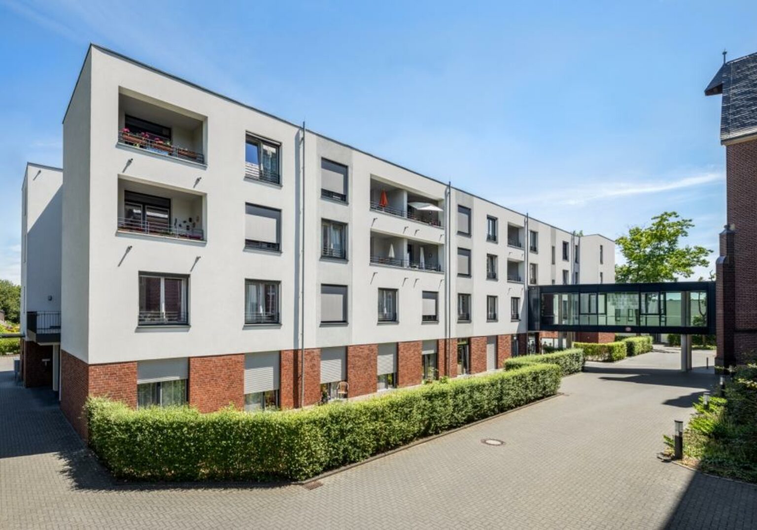 300319_Irmgardisstift_1_klein-1536x1075.2-c-default Moderne Wohngebäude mit Verbindungsbrücke, umgeben von grünen Hecken bei klarem Himmel.