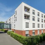 Moderne Wohnanlage mit weißen Fassaden und Balkonen an einem sonnigen Tag, umgeben von gepflegten Hecken.