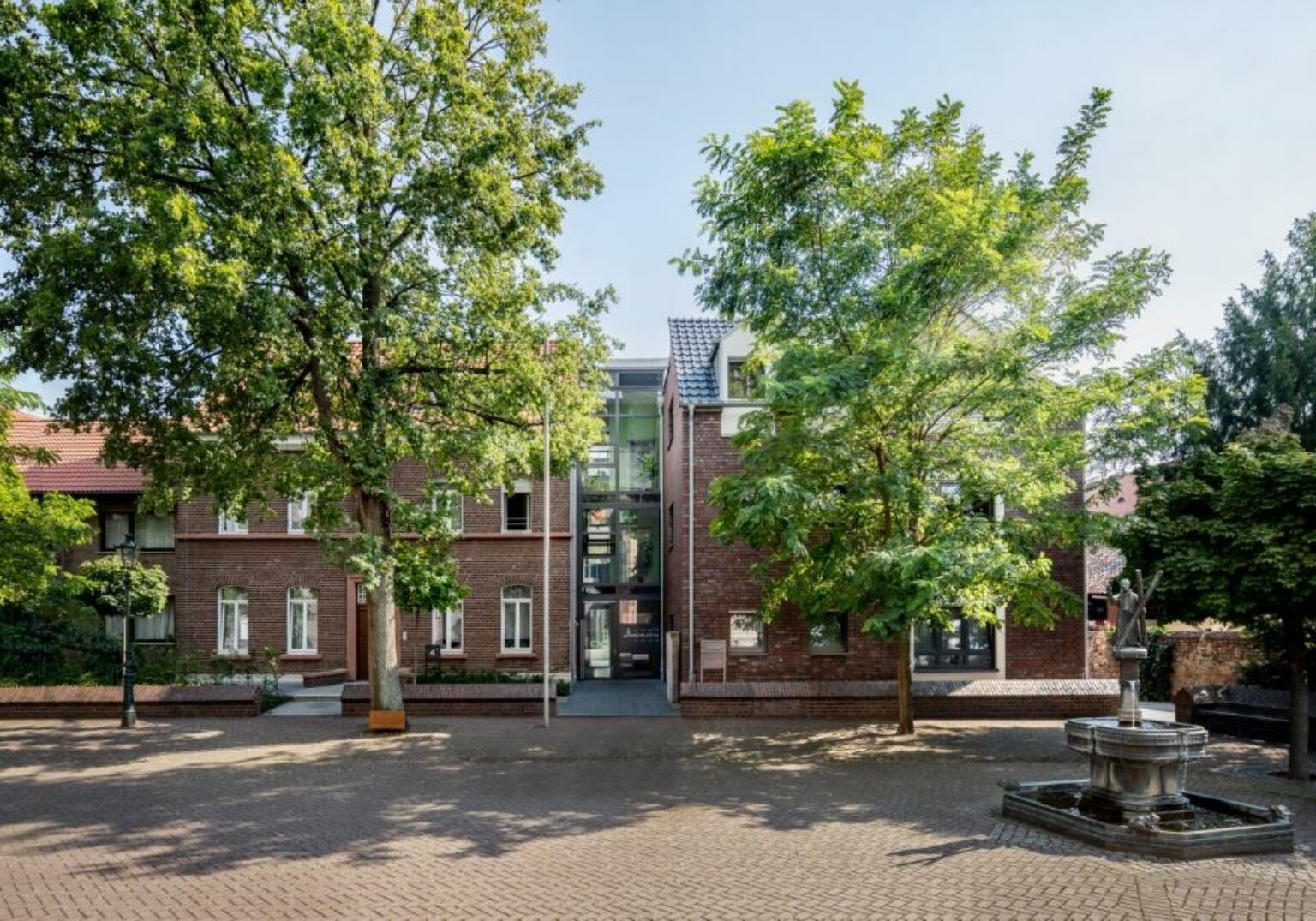Historisches Backsteingebäude mit Bäumen und Springbrunnen auf einem sonnigen Platz dargestellt.