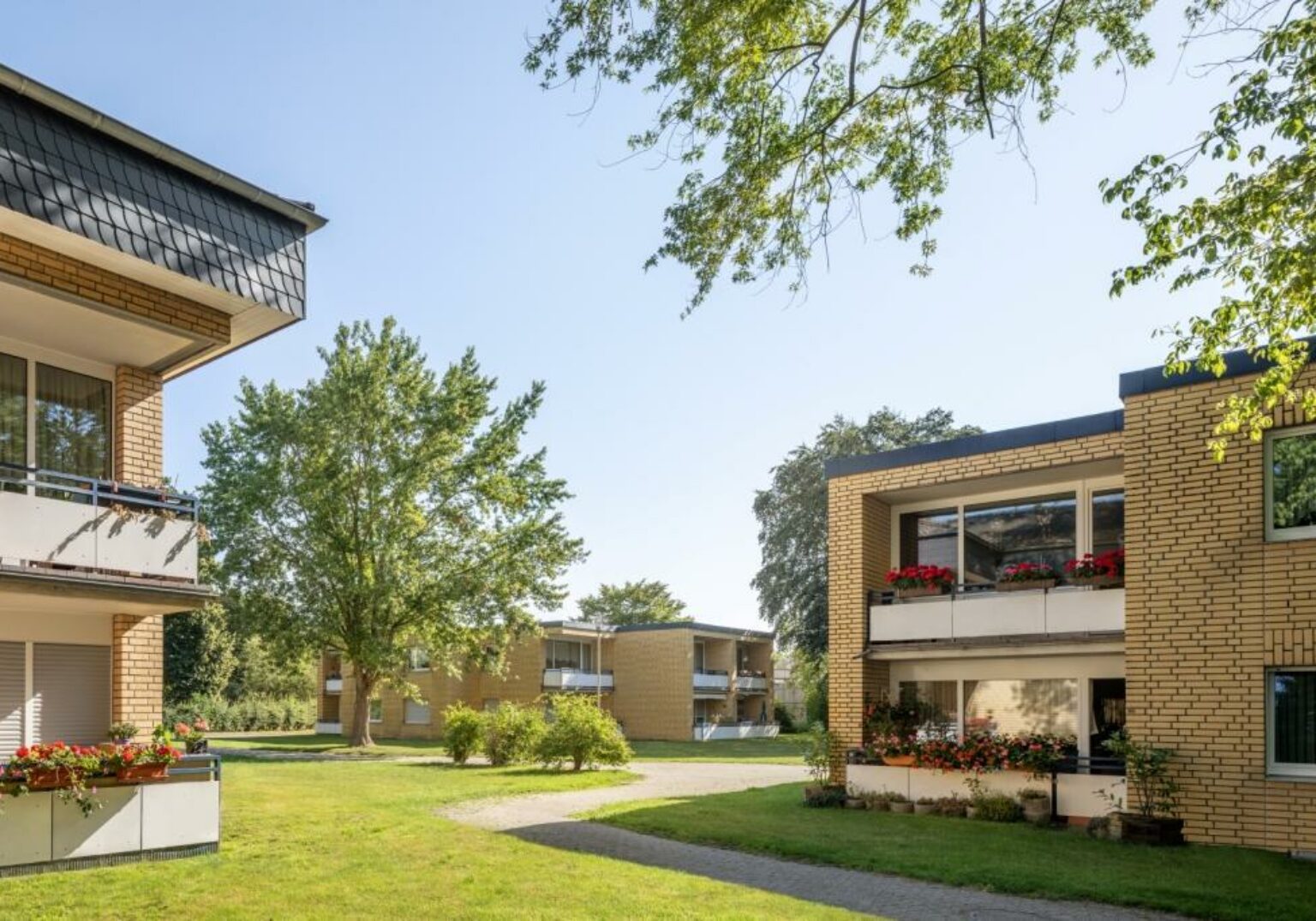 Moderne Wohngebäude mit gepflegten Gärten und Balkonen, umgeben von Bäumen an einem sonnigen Tag.