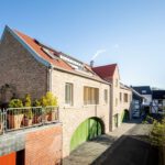 Modernes Backsteinhaus mit roten Dächern und grünen Toren in sonniger Wohngegend. Harmonische Architektur und Natur.