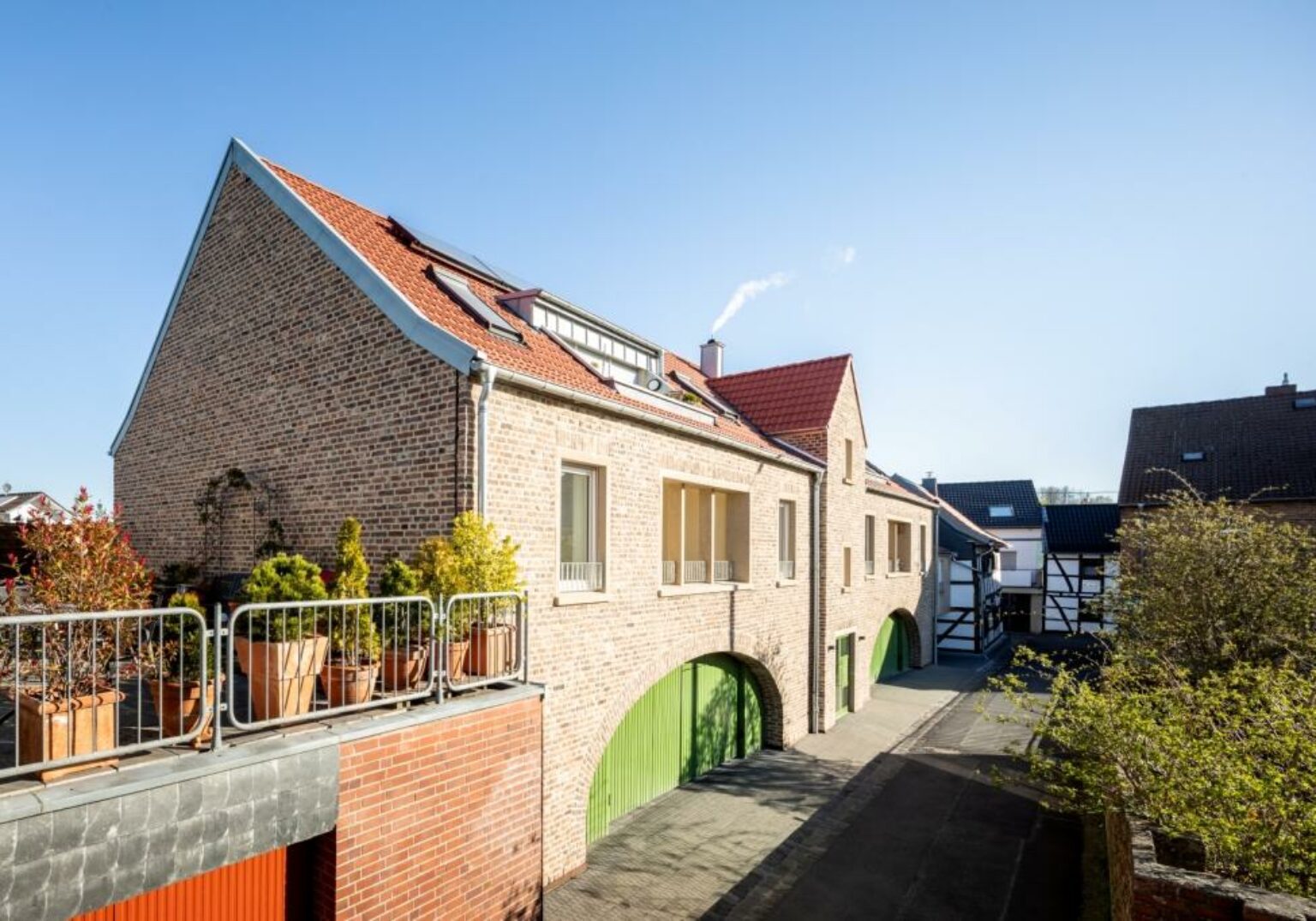 Modernes Backsteinhaus mit roten Dächern und grünen Toren in sonniger Wohngegend. Harmonische Architektur und Natur.