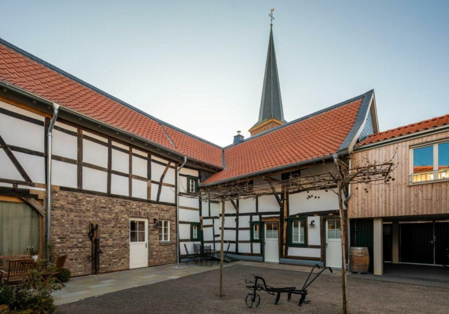 300425_Kirchstrasse_3_klein-1536x1075.2-c-default Fachwerkhaus mit historischer Architektur und Innenhof, im Hintergrund ein Kirchturm bei klarem Himmel.
