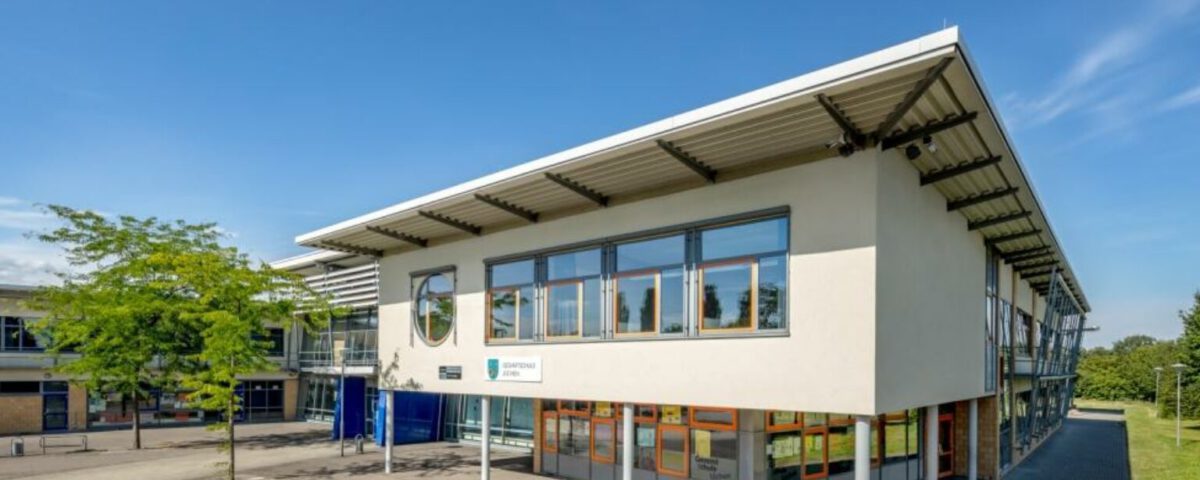 Modernes Schulgebäude mit hellen Fenstern und blauem Himmel im Hintergrund, umgeben von Grünflächen.