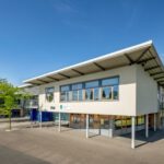 Modernes Schulgebäude mit hellen Fenstern und blauem Himmel im Hintergrund, umgeben von Grünflächen.