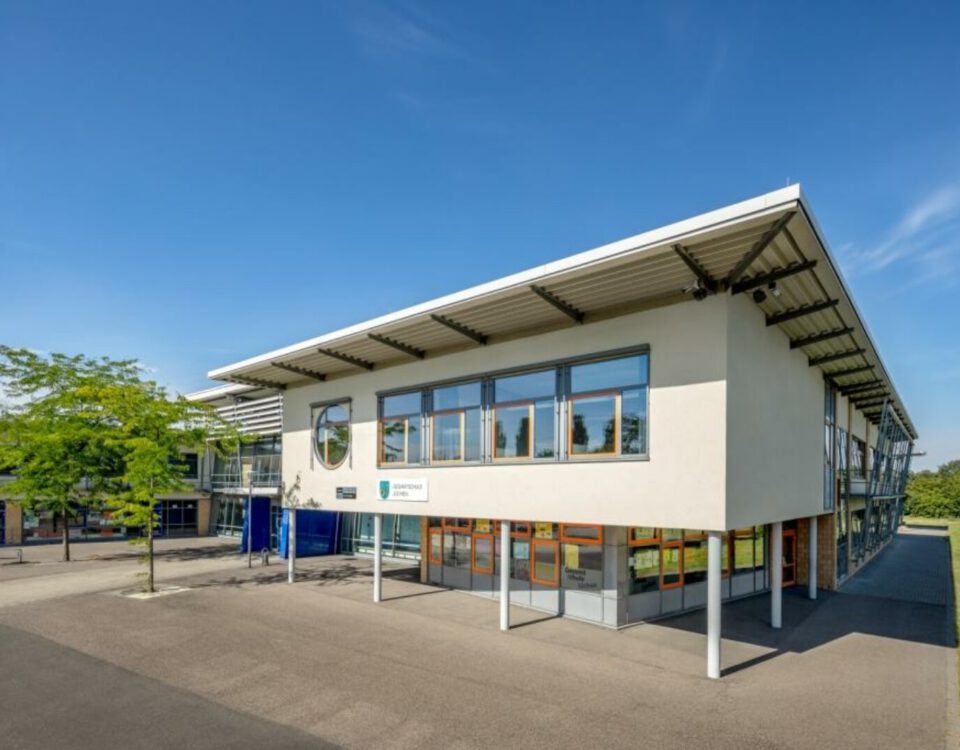 Modernes Schulgebäude mit hellen Fenstern und blauem Himmel im Hintergrund, umgeben von Grünflächen.