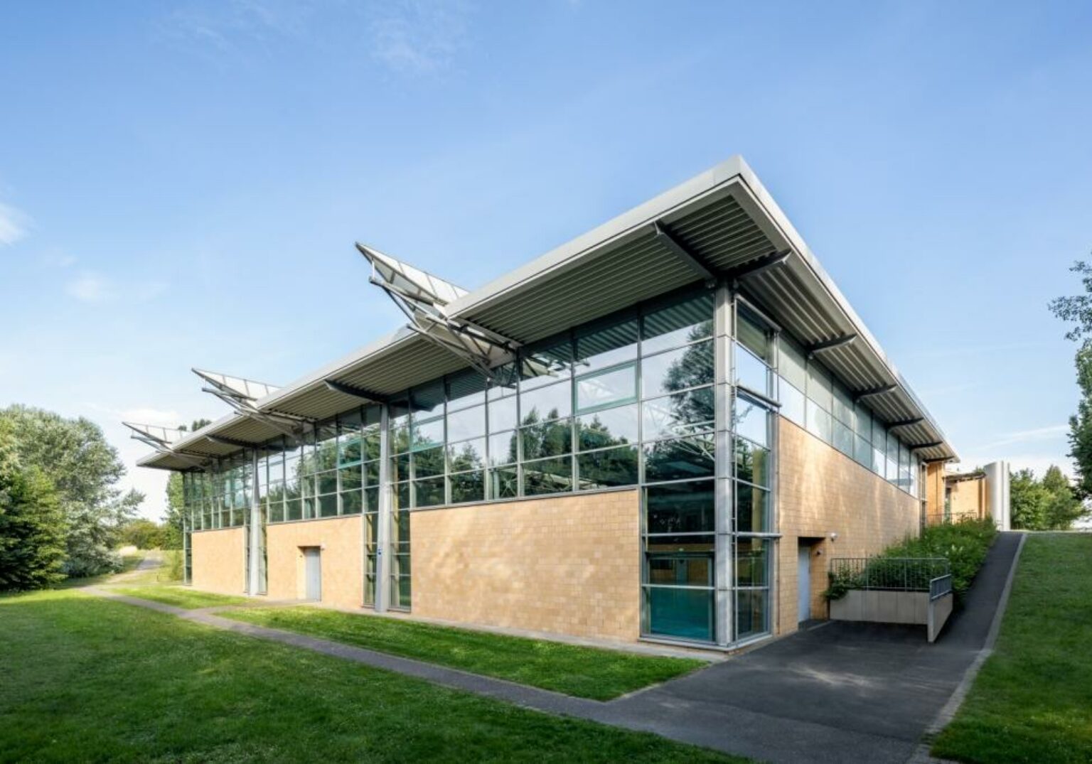 Modernes Gebäude mit Glasfassade und Solarpanelen, umgeben von grünem Rasen und Bäumen, unter blauem Himmel.