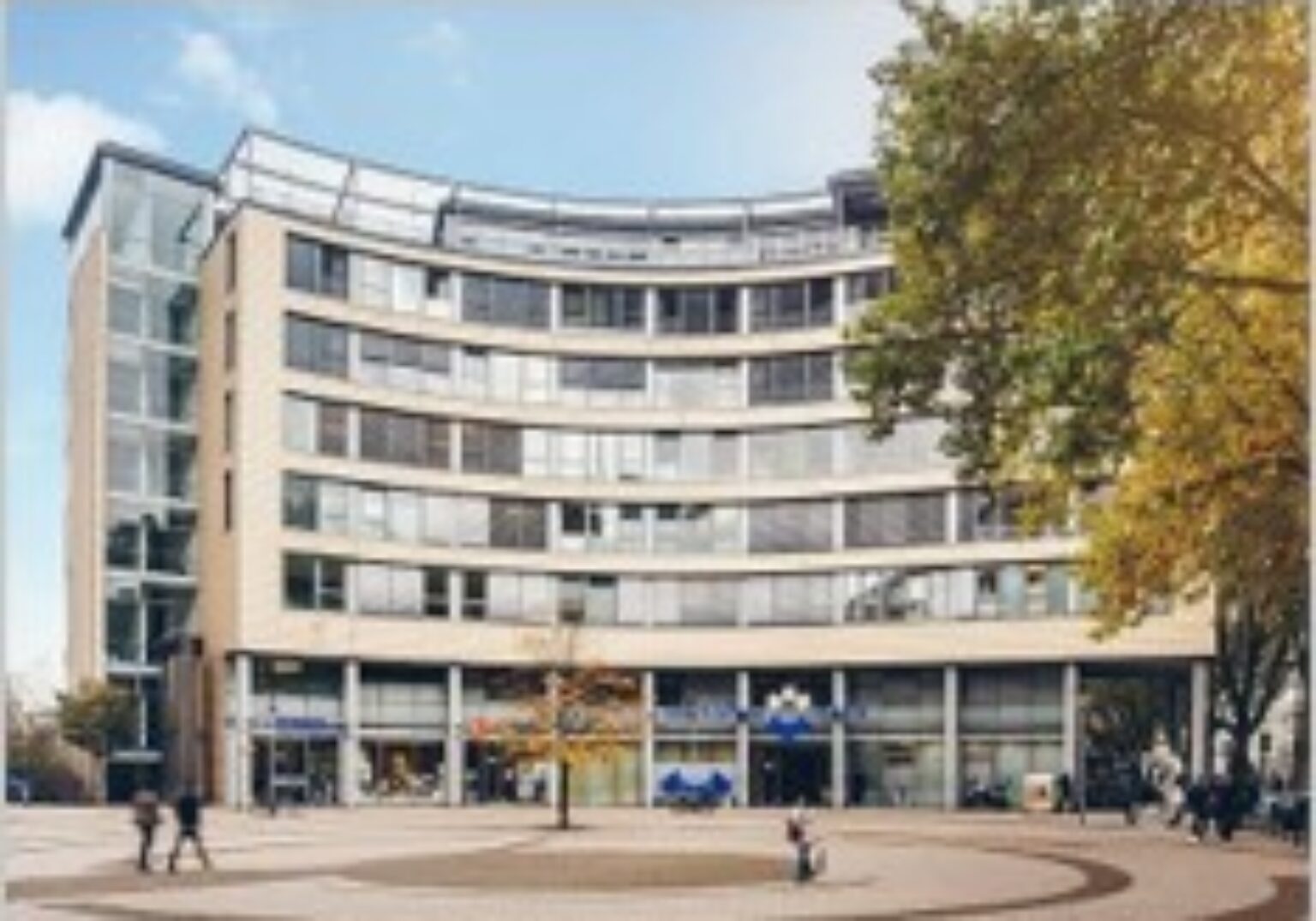 Moderne Glasfassade eines Bürogebäudes mit geschwungener Architektur, umgeben von Bäumen und Passanten.