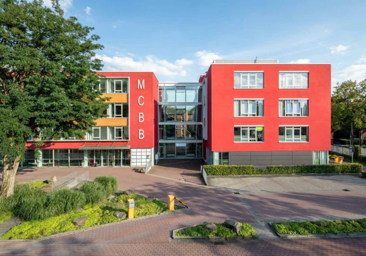 Modernes Gebäude mit roter Fassade und großem Baum im Vordergrund, klare Glasfenster, sonniger Tag.