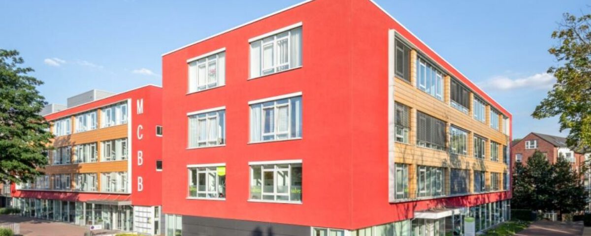 Modernes Bürogebäude mit roter Fassade und großen Fenstern bei Sonnenschein, umgeben von Bäumen und gepflegtem Gehweg.
