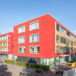 Modernes Bürogebäude mit roter Fassade und großen Fenstern bei Sonnenschein, umgeben von Bäumen und gepflegtem Gehweg.