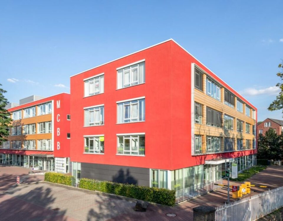 Modernes Bürogebäude mit roter Fassade und großen Fenstern bei Sonnenschein, umgeben von Bäumen und gepflegtem Gehweg.