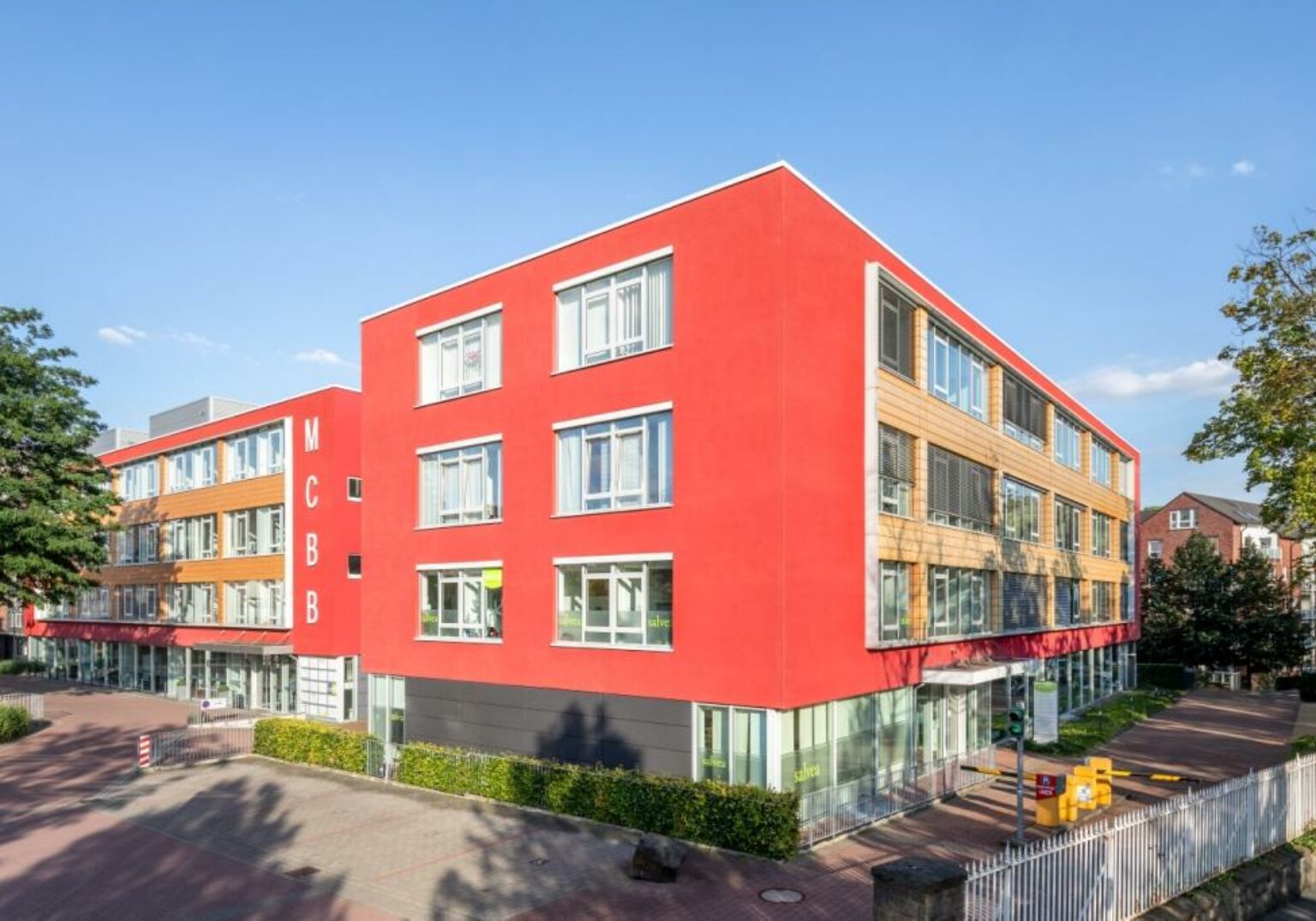 Modernes Bürogebäude mit roter Fassade und großen Fenstern bei Sonnenschein, umgeben von Bäumen und gepflegtem Gehweg.
